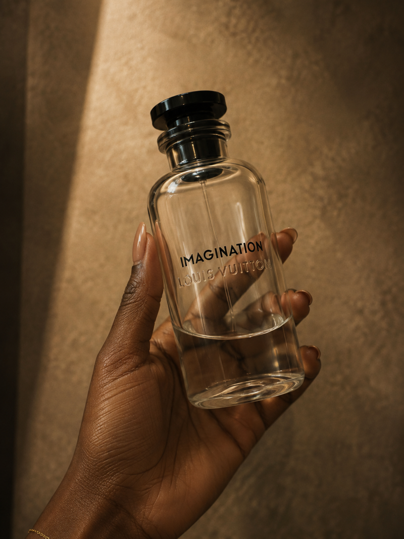 A hand holds a clear glass perfume bottle with a black cap. The bottle is embossed with the text 'IMAGINATION LOUIS VUITTON'.